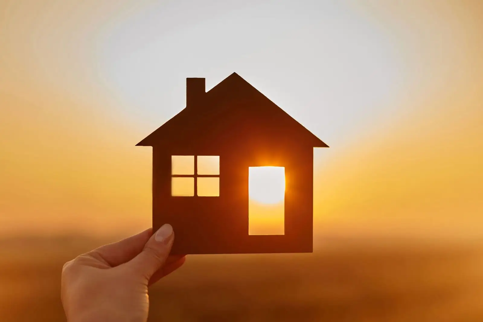 Hand holding a small house cutout against a sunset sky